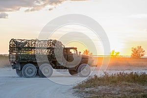 A military car is driving along the sandy road at sunset