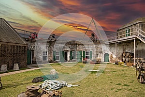 A military fort with red brick walls and green doors with cannons, brown wooden barrels and benches, spools of rope and boat oars