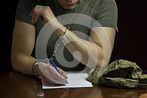 A military man sits at a desk, handcuffed, signing a document on white paper in a dimly lit room.