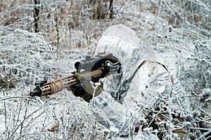 Military man in white camouflage uniform with hood and machinegun in the long winter grass. Soldier stood on knelt and aims of the photo