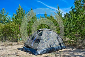 A military tent in an evergreen forest. Tourist and hiker layover on white sand in a small camouflage tent