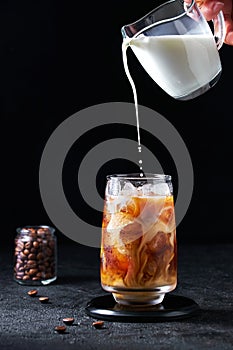 Milk Being Poured into Iced Coffee in Tall Glass on Dark Background. Concept Refreshing Summer Drink