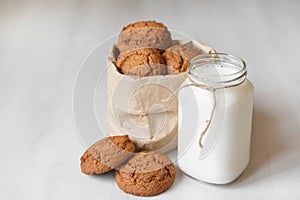 Milk in a glass jar and oatmeal cookies in a paper bag on a white table background. Copy, empty space for text