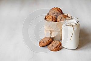 Milk in a glass jar and oatmeal cookies in a paper bag on a white table background. Copy, empty space for text