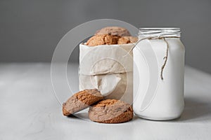 Milk in a glass jar and oatmeal cookies in a paper bag on a white table background. Copy, empty space for text