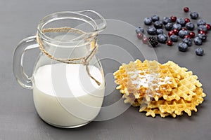 Milk in glass jug. Waffles and berries on table