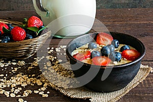 Milk in a jug and oatmeal porridge in a pottery bowl with fresh ripe berries in a wicker bowl