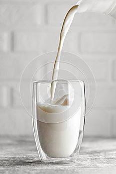 milk pouring in glass from jug on table with white brick wall as background