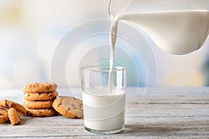 Milk pouring from jug into glass with cookies on wooden table