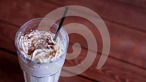 Milkshake in high glass on wooden table. Cold beverage with topping of whipped cream and chocolate powder on brown background