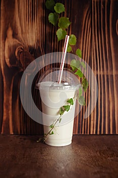 Milkshake in a plastic glass with a straw stands on a wooden background. Liquid ice cream helps you cool off in the summer heat.