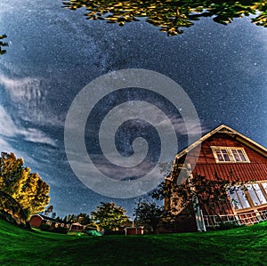 Milky Way above the Swedish traditional wooden red house at the left side of night scene in garden. Starry sky, green grass