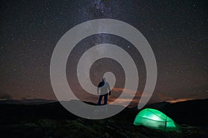 Milky way and starry sky over night scene outdoors in the forest and the mountains with green tent and silhouette of man infront.