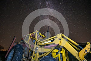 Milky way and starry sky over night scene outdoors, yellow old and rustic tractor used as foreground.