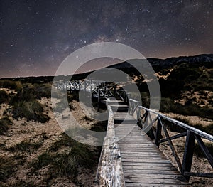 Milky way starscape above wood board walk pathway leading across sand dunes with grass