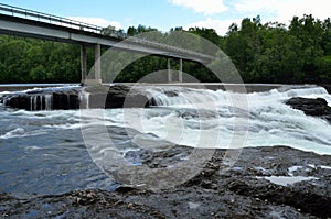 Milky white massive long waterfall down slippery valley rocks and stones in summer with car bridge