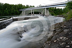 Milky white massive long waterfall down slippery valley rocks and stones in summer with car bridge