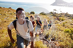 Millennial African American man leading friends hiking single file uphill on a path by the coast