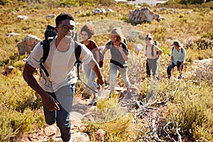 Millennial African American man leading friends hiking single file uphill on a path in countryside