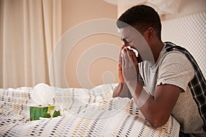 Millennial African American man sitting up in bed blowing nose, side view, close up