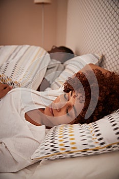 Millennial African American woman lying asleep in bed, her partner in the background, vertical