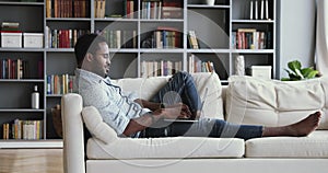 Millennial african man sit in livingroom on sofa using laptop