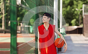 Millennial Asian basketball player with ball standing near fence at outdoor sports arena