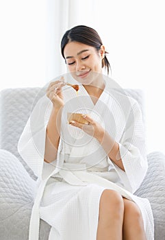 Millennial Asian beautiful pretty cheerful female model in white clean bathrobe sitting smiling on chair holding honey sugar wax