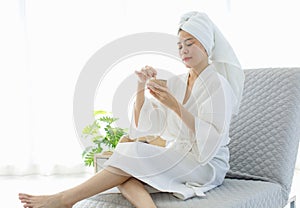 Millennial Asian beautiful pretty cheerful female model in white clean bathrobe sitting smiling on chair holding honey sugar wax