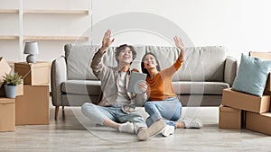 Millennial Asian couple sitting on floor among carton boxes, using digital tablet, dreaming about design of new home