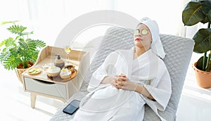 Millennial Asian female customer in white clean bathrobe and towel sitting on armchair closed eyes with cucumbers waiting for