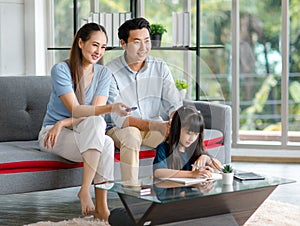 Millennial Asian happy family father and mother sitting on cozy sofa couch holding remote watching television while little young
