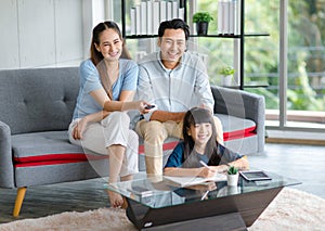 Millennial Asian happy family father and mother sitting on cozy sofa couch holding remote watching television while little young