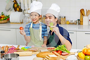 Millennial Asian little young boy chef wearing tall white cook hat and apron standing smiling holding sliced tomato posing