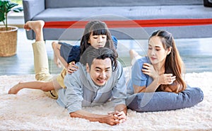 Millennial Asian lovely happy family father mother laying down on carpet floor while young daughter girl sitting piggy back on dad