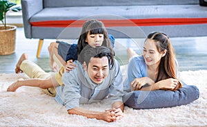 Millennial Asian lovely happy family father mother laying down on carpet floor while young daughter girl sitting piggy back on dad