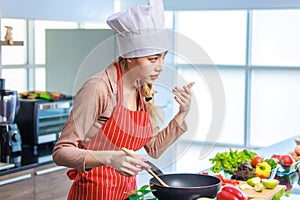 Millennial Asian young cheerful female housewife chef wearing white tall cook hat and apron standing smiling waving hand smelling