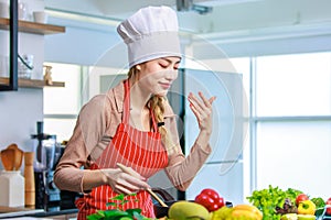 Millennial Asian young cheerful female housewife chef wearing white tall cook hat and apron standing smiling waving hand smelling