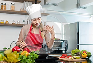 Millennial Asian young cheerful female housewife chef wearing white tall cook hat and apron standing smiling waving hand smelling