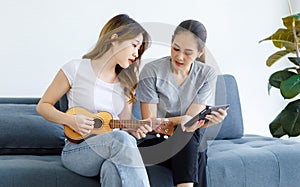 Millennial Asian young happy female lesbian LGBTQ lover couple sitting together on sofa playing small ukulele in living room