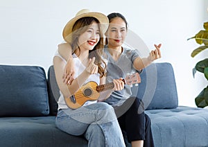 Millennial Asian young happy female lesbian LGBTQ lover couple sitting together on sofa playing small ukulele in living room