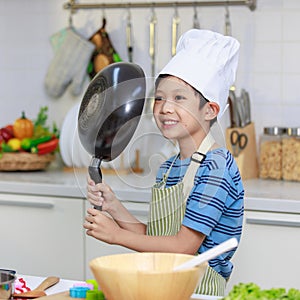 Millennial Asian young little boy chef wearing white tall cook hat and apron standing smiling holding cooking pan posing taking