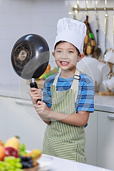 Millennial Asian young little boy chef wearing white tall cook hat and apron standing smiling holding cooking pan posing taking