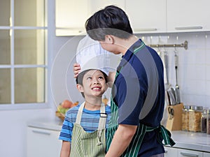 Millennial Asian young little boy pastry chef wearing white tall cook hat and apron standing smiling posing together with father