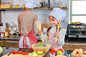 Millennial Asian young little cute girl chef daughter with white tall cook hat and apron standing smiling holding wooden spoon and
