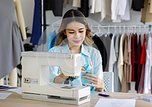 Millennial Asian young professional female designer dressmaker businesswoman with measuring tape around neck sitting using sewing