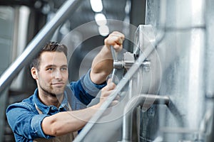 Millennial attractive male worker or owner in apron turns valve on big boiler for beer in interior of plant