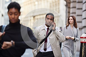 Millennial black businessman walking in a busy London street using smartphone, selective focus photo