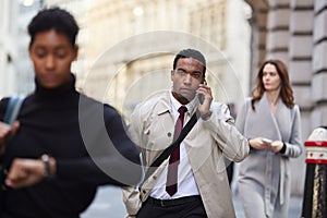 Millennial black businessman walking in a busy London street using smartphone, selective focus