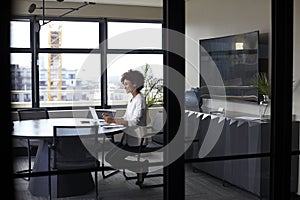 Millennial black businesswoman working alone in an office meeting room, seen through glass wall